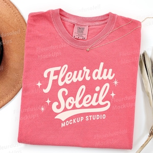 May include: A coral-colored short-sleeve t-shirt with a crew neckline and a Comfort Colors brand label. The front features the white text Fleur du Soleil MOCKUP STUDIO in a cursive font, accented by small four-pointed star graphics.