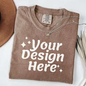 Peut inclure: Un t-shirt marron avec le texte "Your Design Here" en blanc. Le t-shirt a un col rond et une petite &eacute;tiquette. Un collier argent&eacute; avec un pendentif est sur le t-shirt. Un chapeau marron et des feuilles s&eacute;ch&eacute;es sont en arri&egrave;re-plan.