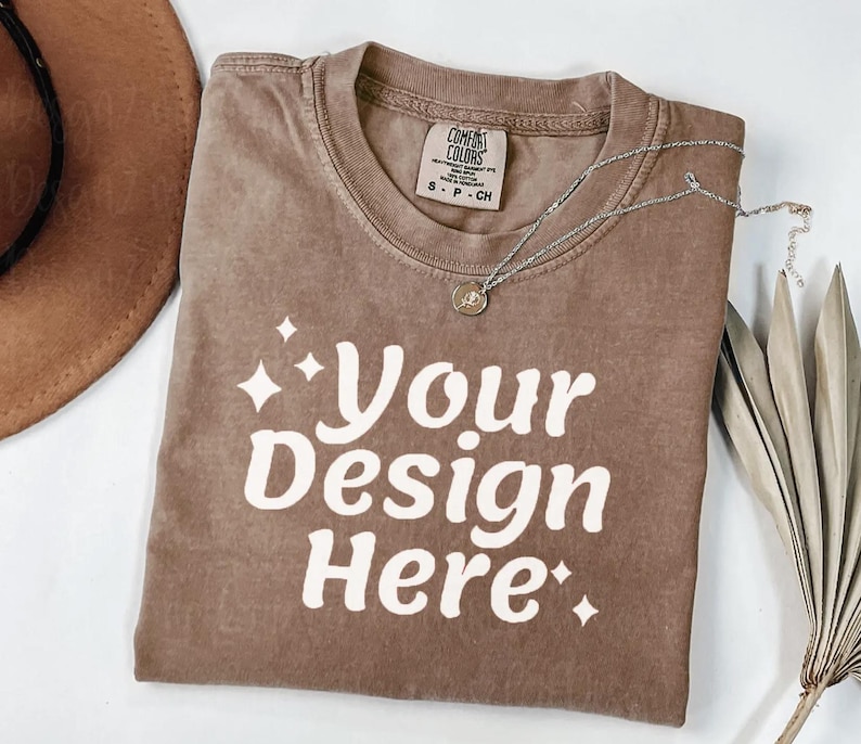 Peut inclure: Un t-shirt marron avec le texte "Your Design Here" en blanc. Le t-shirt a un col rond et une petite &eacute;tiquette. Un collier argent&eacute; avec un pendentif est sur le t-shirt. Un chapeau marron et des feuilles s&eacute;ch&eacute;es sont en arri&egrave;re-plan.