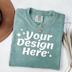 Peut inclure: Un t-shirt vert d'eau avec le texte "Your Design Here" en blanc, avec des accents d'&eacute;clats d'&eacute;toiles. Un collier argent&eacute; avec un pendentif repose sur le t-shirt. Un chapeau marron et une plante s&eacute;ch&eacute;e sont en arri&egrave;re-plan.