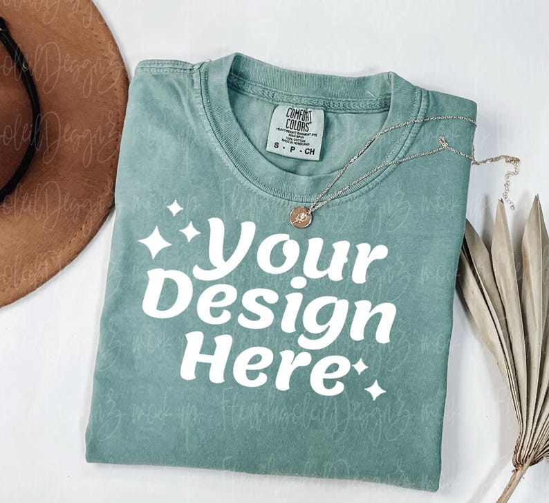 Peut inclure: Un t-shirt vert d'eau avec le texte "Your Design Here" en blanc, avec des accents d'&eacute;clats d'&eacute;toiles. Un collier argent&eacute; avec un pendentif repose sur le t-shirt. Un chapeau marron et une plante s&eacute;ch&eacute;e sont en arri&egrave;re-plan.