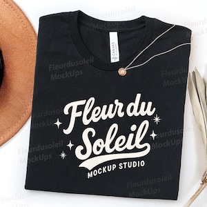 May include: Black t-shirt with the text "Fleur du Soleil MOCKUP STUDIO" in white script. A gold necklace with a circular pendant rests on the shirt. A brown hat and dried plant are in the background.