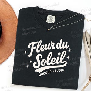 May include: Black t-shirt with the text "Fleur du Soleil" in white script. The shirt has a crew neck and short sleeves. A gold necklace with a star pendant and a brown hat are also visible.