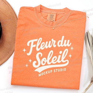 Puede incluir: Una camiseta naranja con el texto "Fleur du Soleil" en escritura blanca. La camiseta tiene cuello redondo y mangas cortas. Un collar dorado con un colgante de estrella está sobre la camiseta. Un sombrero marrón es visible.