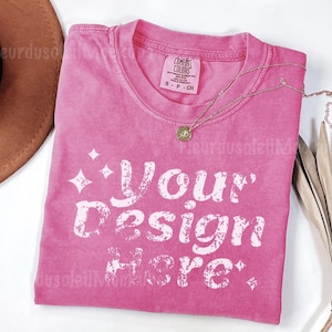 Peut inclure: Un t-shirt rose avec le texte "Your Design Here" en blanc, avec un collier dor&eacute; et un chapeau marron. Le t-shirt est de la marque Comfort Colors, taille S-CH.