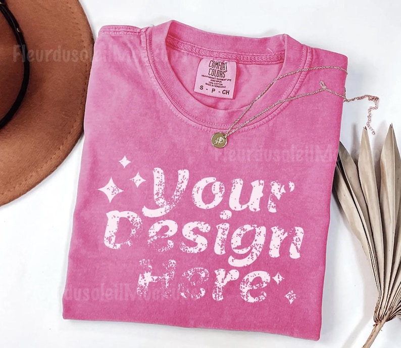 Peut inclure: Un t-shirt rose avec le texte "Your Design Here" en blanc, avec un collier dor&eacute; et un chapeau marron. Le t-shirt est de la marque Comfort Colors, taille S-CH.