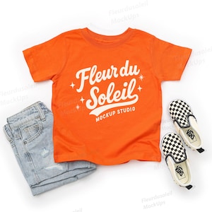 May include: An orange t-shirt with the text "Fleur du Soleil MOCKUP STUDIO" in white, paired with light blue denim jeans and black and white checkered slip-on shoes.