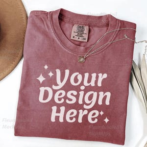 Peut inclure: Un t-shirt rose poussi&eacute;reux avec le texte "Your Design Here" en blanc. Un collier argent&eacute; avec un pendentif repose sur le t-shirt. Un chapeau marron et une plante s&eacute;ch&eacute;e sont en arri&egrave;re-plan.