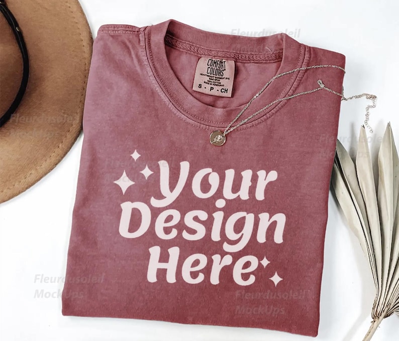 Peut inclure: Un t-shirt rose poussi&eacute;reux avec le texte "Your Design Here" en blanc. Un collier argent&eacute; avec un pendentif repose sur le t-shirt. Un chapeau marron et une plante s&eacute;ch&eacute;e sont en arri&egrave;re-plan.