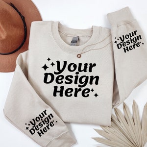 May include: A beige sweatshirt with the text "Your Design Here" on the front and both sleeves. The sweatshirt is laid flat on a white surface with a brown hat and a dried palm leaf.