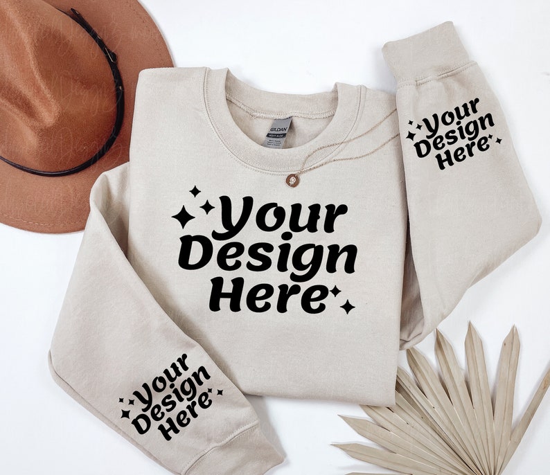 May include: A beige sweatshirt with the text "Your Design Here" on the front and both sleeves. The sweatshirt is laid flat on a white surface with a brown hat and a dried palm leaf.