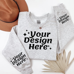 May include: A grey sweatshirt with the text "Your Design Here" on the front and both sleeves. The sweatshirt is laid flat on a white surface with a brown hat and a dried palm leaf.