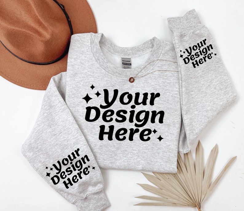 May include: A grey sweatshirt with the text "Your Design Here" on the front and both sleeves. The sweatshirt is laid flat on a white surface with a brown hat and a dried palm leaf.