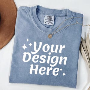 Peut inclure: Un t-shirt bleu Comfort Colors avec le texte "Your Design Here" en blanc. Un collier en or avec un pendentif est drap&eacute; sur le t-shirt. Le t-shirt est pli&eacute; et pos&eacute; sur une surface blanche.