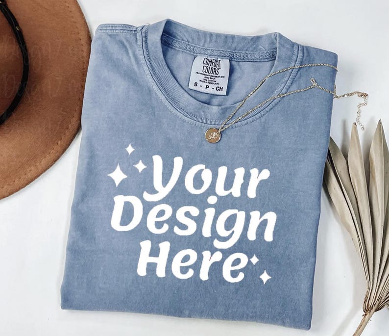 Peut inclure: Un t-shirt bleu Comfort Colors avec le texte "Your Design Here" en blanc. Un collier en or avec un pendentif est drap&eacute; sur le t-shirt. Le t-shirt est pli&eacute; et pos&eacute; sur une surface blanche.