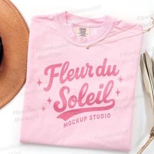 May include: A light pink t-shirt with the words "Fleur du Soleil" in a cursive font. The shirt has a crew neck and a small tag on the collar. A gold necklace and a brown hat are also visible.