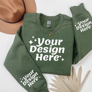 May include: A green sweatshirt with the text "Your Design Here" on the front and both sleeves. The sweatshirt is laid flat on a white surface with a brown hat and a dried palm leaf.