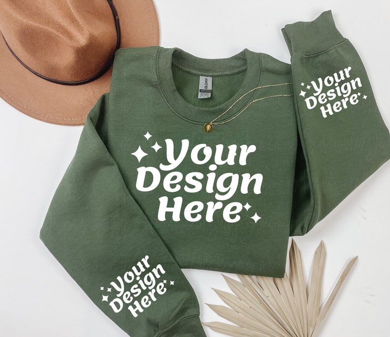 May include: A green sweatshirt with the text "Your Design Here" on the front and both sleeves. The sweatshirt is laid flat on a white surface with a brown hat and a dried palm leaf.