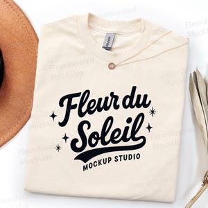 Könnte beinhalten: Cremefarbenes T-Shirt mit dem Text "Fleur du Soleil Mockup Studio" in schwarzer Schrift. Eine goldene Halskette liegt auf dem Shirt. Ein brauner Hut und eine getrocknete Pflanze sind im Hintergrund.