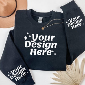 May include: Black sweatshirt with white text that says "Your Design Here" on the front and both sleeves. The sweatshirt is laid flat on a white surface with a brown hat and a dried palm leaf.