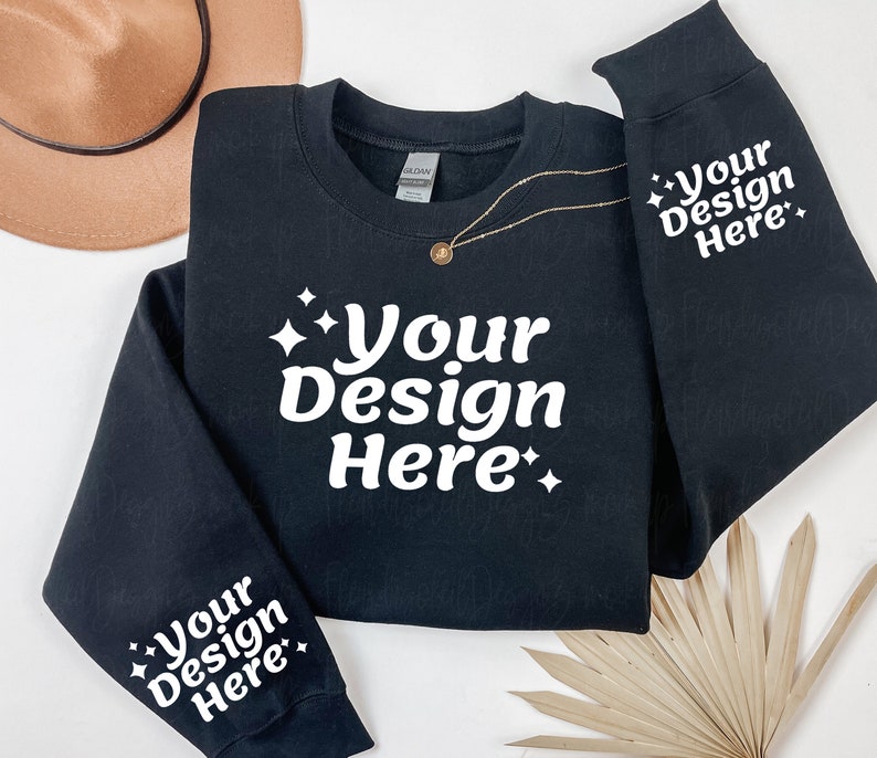 May include: Black sweatshirt with white text that says "Your Design Here" on the front and both sleeves. The sweatshirt is laid flat on a white surface with a brown hat and a dried palm leaf.