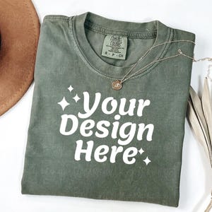 Peut inclure: Un t-shirt vert olive pli&eacute; avec le texte "Your Design Here" en blanc. Un collier dor&eacute; avec un pendentif repose sur le t-shirt. L'&eacute;tiquette du t-shirt indique "Comfort Colors". Un chapeau marron est visible en haut &agrave; gauche.