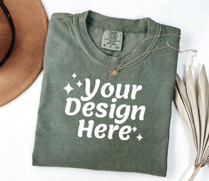Peut inclure: Un t-shirt vert olive pli&eacute; avec le texte "Your Design Here" en blanc. Un collier dor&eacute; avec un pendentif repose sur le t-shirt. L'&eacute;tiquette du t-shirt indique "Comfort Colors". Un chapeau marron est visible en haut &agrave; gauche.