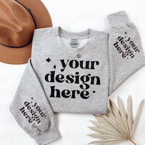 May include: A grey sweatshirt with the text "your design here" printed in black on the front and sleeves. The sweatshirt is laid flat on a white surface with a brown hat and a dried palm leaf.