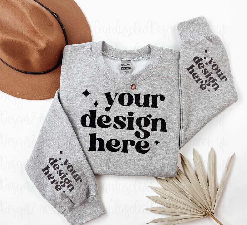 May include: A grey sweatshirt with the text "your design here" printed in black on the front and sleeves. The sweatshirt is laid flat on a white surface with a brown hat and a dried palm leaf.