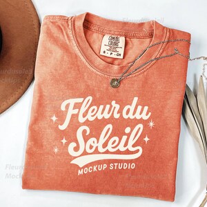 Peut inclure: Un t-shirt orange Comfort Colors avec le texte "Fleur du Soleil" en &eacute;criture blanche. Un collier en argent avec un petit pendentif repose sur le t-shirt. Un chapeau marron et des feuilles de palmier s&eacute;ch&eacute;es sont &eacute;galement dans l'image.