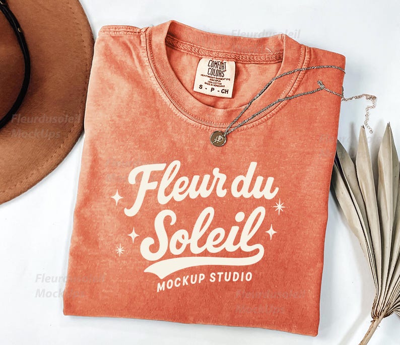 Peut inclure: Un t-shirt orange Comfort Colors avec le texte "Fleur du Soleil" en &eacute;criture blanche. Un collier en argent avec un petit pendentif repose sur le t-shirt. Un chapeau marron et des feuilles de palmier s&eacute;ch&eacute;es sont &eacute;galement dans l'image.