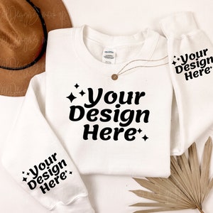 May include: White sweatshirt with the text "Your Design Here" printed on the front and sleeves. The sweatshirt is laid flat on a white surface with a brown hat and a dried palm leaf.