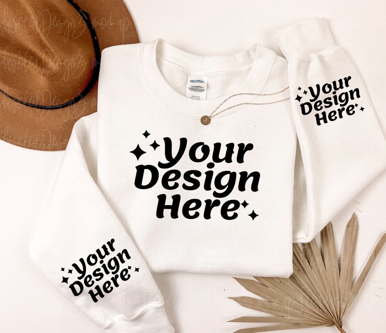 May include: White sweatshirt with the text "Your Design Here" printed on the front and sleeves. The sweatshirt is laid flat on a white surface with a brown hat and a dried palm leaf.