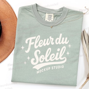 May include: A light green t-shirt with the text "Fleur du Soleil" in white script. The shirt has a crew neck and short sleeves. A gold necklace with a star pendant and a brown hat are also in the image.