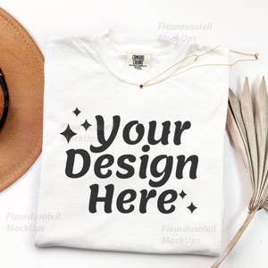 Puede incluir: Una camiseta blanca con el texto "Your Design Here" en negrita y en color negro. La camiseta está doblada y tiene una etiqueta Comfort Colors. También hay un collar dorado con un colgante de estrella y un sombrero marrón.