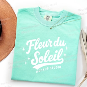 May include: A light teal t-shirt with the text "Fleur du Soleil" in white script. The shirt has a crew neck and a small tag that says "Comfort Colors". A gold necklace with a star pendant is on the shirt.