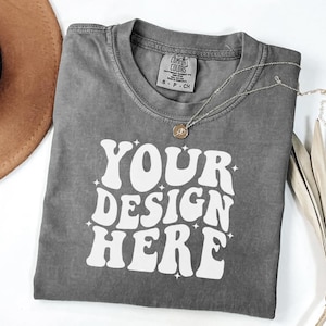 Peut inclure: Un t-shirt gris Comfort Colors avec le texte "YOUR DESIGN HERE" en blanc, un collier argent&eacute; et un chapeau marron. Le t-shirt a un col rond et une petite &eacute;tiquette. Le design est dans une police r&eacute;tro.