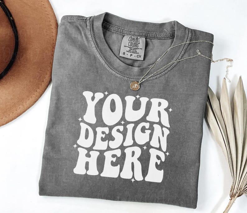 Peut inclure: Un t-shirt gris Comfort Colors avec le texte "YOUR DESIGN HERE" en blanc, un collier argent&eacute; et un chapeau marron. Le t-shirt a un col rond et une petite &eacute;tiquette. Le design est dans une police r&eacute;tro.