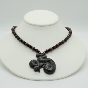 May include: A black stone snake pendant necklace with red and black beads. The snake is curled up and has a detailed design.