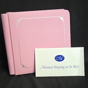 May include: A square, light pink photo album with a silver-colored border design on the front cover. Next to the album is a white rectangular card featuring a blue oval logo with the letter M and the text Memory Keeping at Its Best.