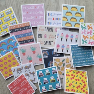 May include: A collection of square greeting cards featuring various colorful patterns including cassette tapes, tacos, rainbows, ice cream cones, balloon animals, pineapples, cherries, camels, typewriters, and text phrases like Let's Cuddle and Best Day Ever.