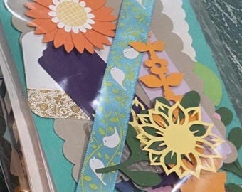 May include: A craft supply kit in a clear plastic sleeve containing various paper die-cuts including an orange flower, a yellow flower, green leaves, and a light blue ribbon with a white bird and vine pattern, layered over teal and tan cardstock.