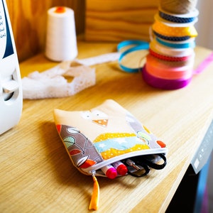 Può includere: Una piccola pochette con cerniera a forma di gufo realizzata in tessuto patchwork con uno schema di colori marrone, beige e arancione. La pochette è aperta e contiene diverse penne e pennarelli.
