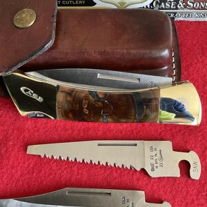 Puede incluir: Una navaja plegable Case & Sons con mango de veta de madera marr&oacute;n y hoja de acero inoxidable. La navaja va acompa&ntilde;ada de dos hojas adicionales, una etiquetada "SAW" y la otra "CLIP". La navaja y las hojas est&aacute;n sobre una superficie roja.