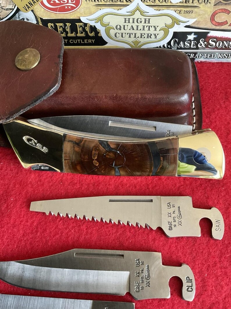 Puede incluir: Una navaja plegable Case & Sons con mango de veta de madera marr&oacute;n y hoja de acero inoxidable. La navaja va acompa&ntilde;ada de dos hojas adicionales, una etiquetada "SAW" y la otra "CLIP". La navaja y las hojas est&aacute;n sobre una superficie roja.