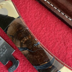 Puede incluir: Primer plano de un cuchillo personalizado con mango de lat&oacute;n e incrustaciones de vetas de madera oscura. La hoja es parcialmente visible, junto con una peque&ntilde;a herramienta de sierra. El cuchillo descansa sobre una superficie roja.