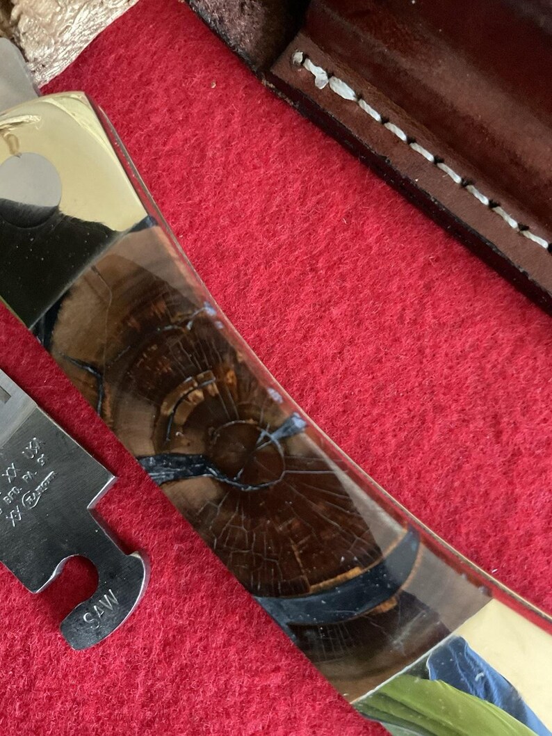 Puede incluir: Primer plano de un cuchillo personalizado con mango de lat&oacute;n e incrustaciones de vetas de madera oscura. La hoja es parcialmente visible, junto con una peque&ntilde;a herramienta de sierra. El cuchillo descansa sobre una superficie roja.