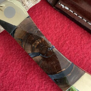 Puede incluir: Primer plano de un mango de cuchillo con un patr&oacute;n de vetas de madera. El mango presenta una combinaci&oacute;n de madera marr&oacute;n clara y oscura, acentuada con secciones negras y doradas. La hoja es parcialmente visible.