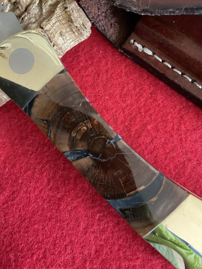 Puede incluir: Primer plano de un mango de cuchillo con un patr&oacute;n de vetas de madera. El mango presenta una combinaci&oacute;n de madera marr&oacute;n clara y oscura, acentuada con secciones negras y doradas. La hoja es parcialmente visible.