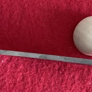 Puede incluir: Primer plano de un objeto esf&eacute;rico blanco y una herramienta met&aacute;lica larga y delgada con un extremo puntiagudo. Los objetos est&aacute;n sobre un fondo rojo.